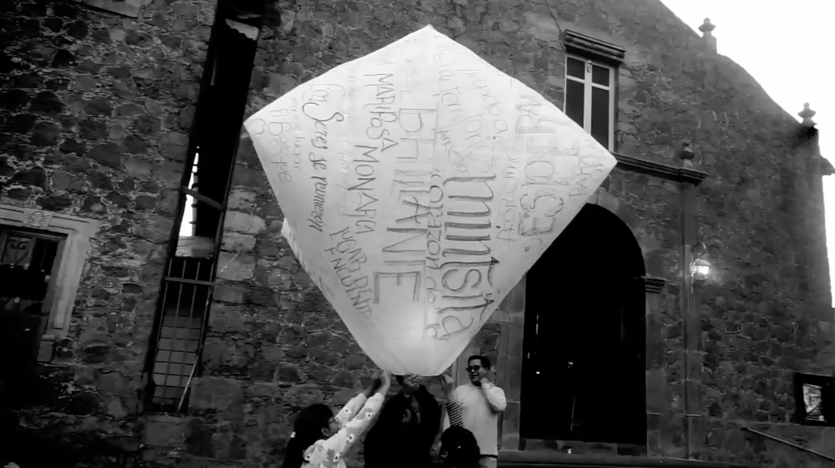 Black and white image of several people holding a large paper lantern above them. The lantern is decorated with various words.